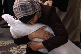 Une mère en larmes tient le corps de son enfant tué, symbolisant la douleur et le chagrin au milieu des conflits. En arrière-plan, une mention des envois d'armes de la France à Israël souligne la contradiction entre les discours de paix et les actions militaires.